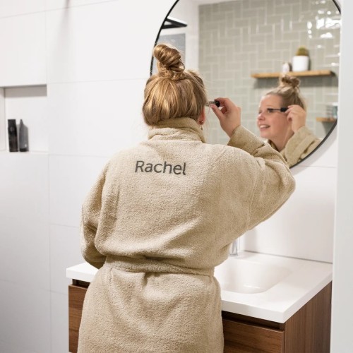 Vrouw die in de spiegel haar make-up doet en een prachtige badjas aan heeft met haar eigen naam achterop. van yoursurprise.nl